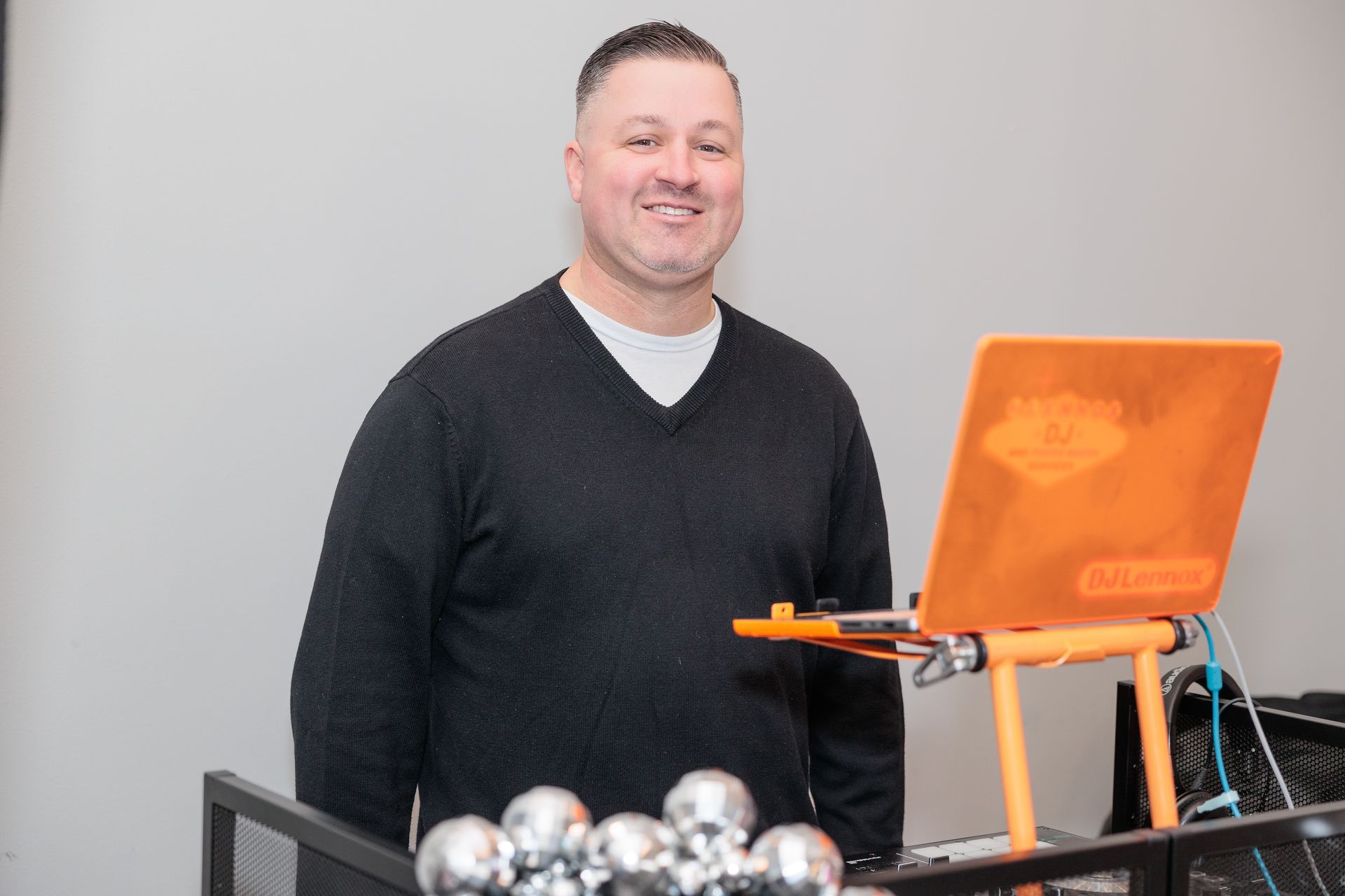 Man in black sweater at DJ setup with orange laptop, smiling.