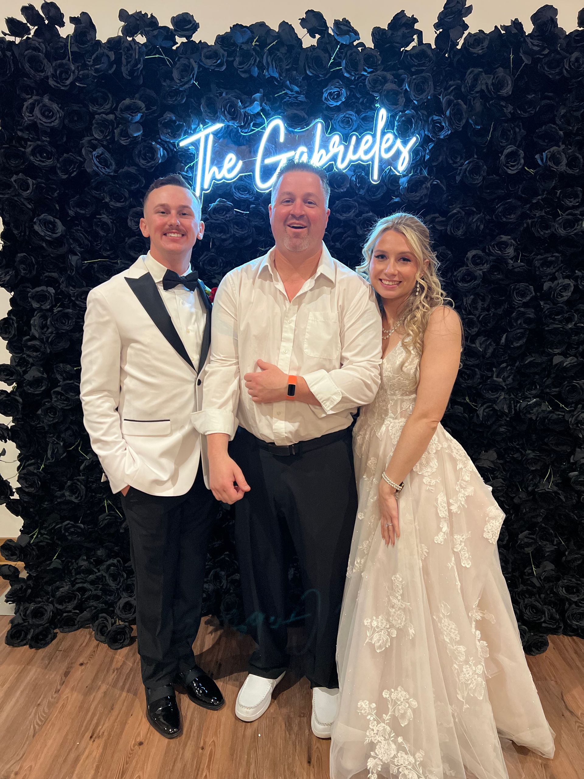 Three people pose in front of a black floral wall, neon sign says 