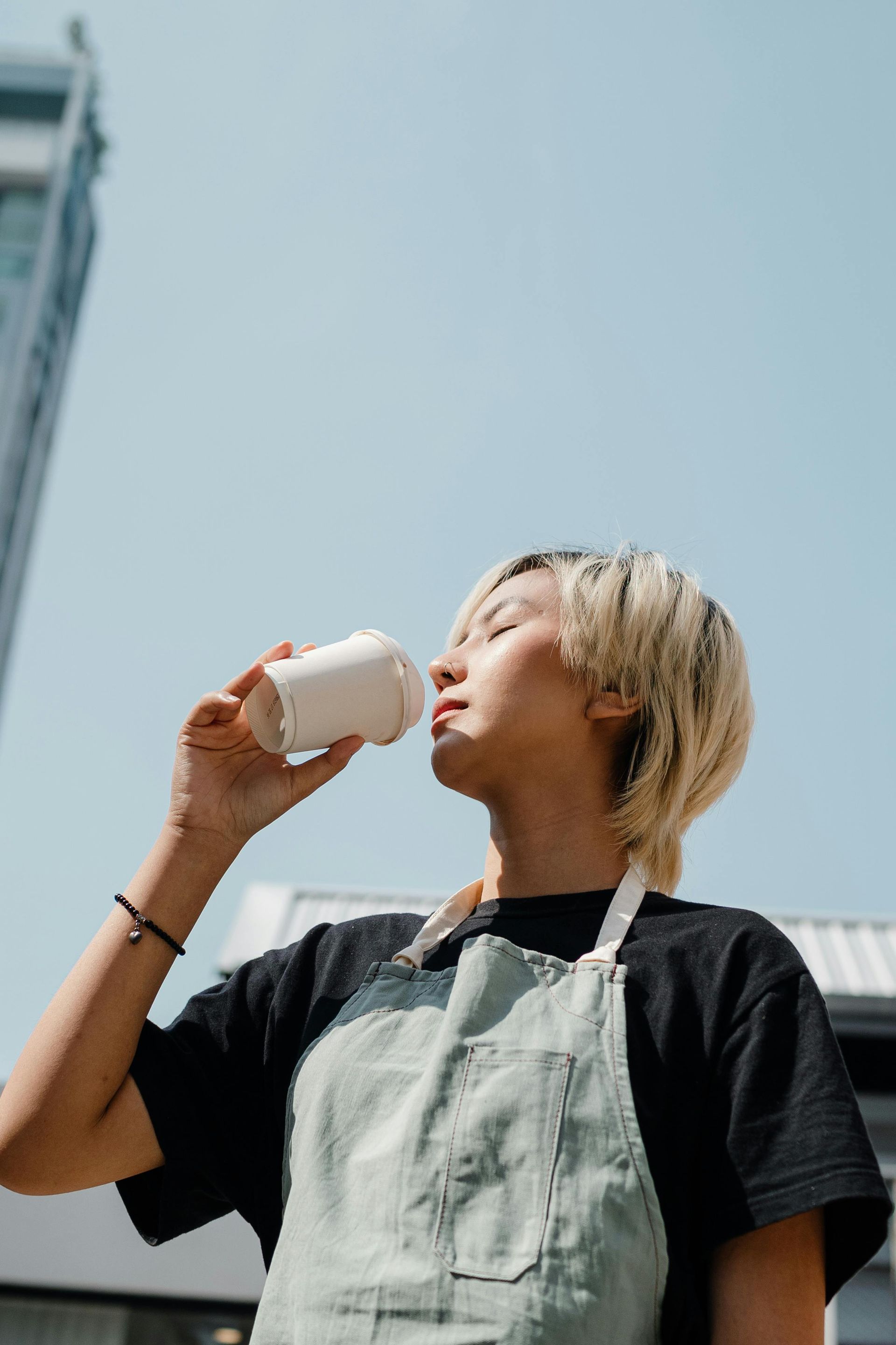 A woman in an apron is drinking from a white cup.