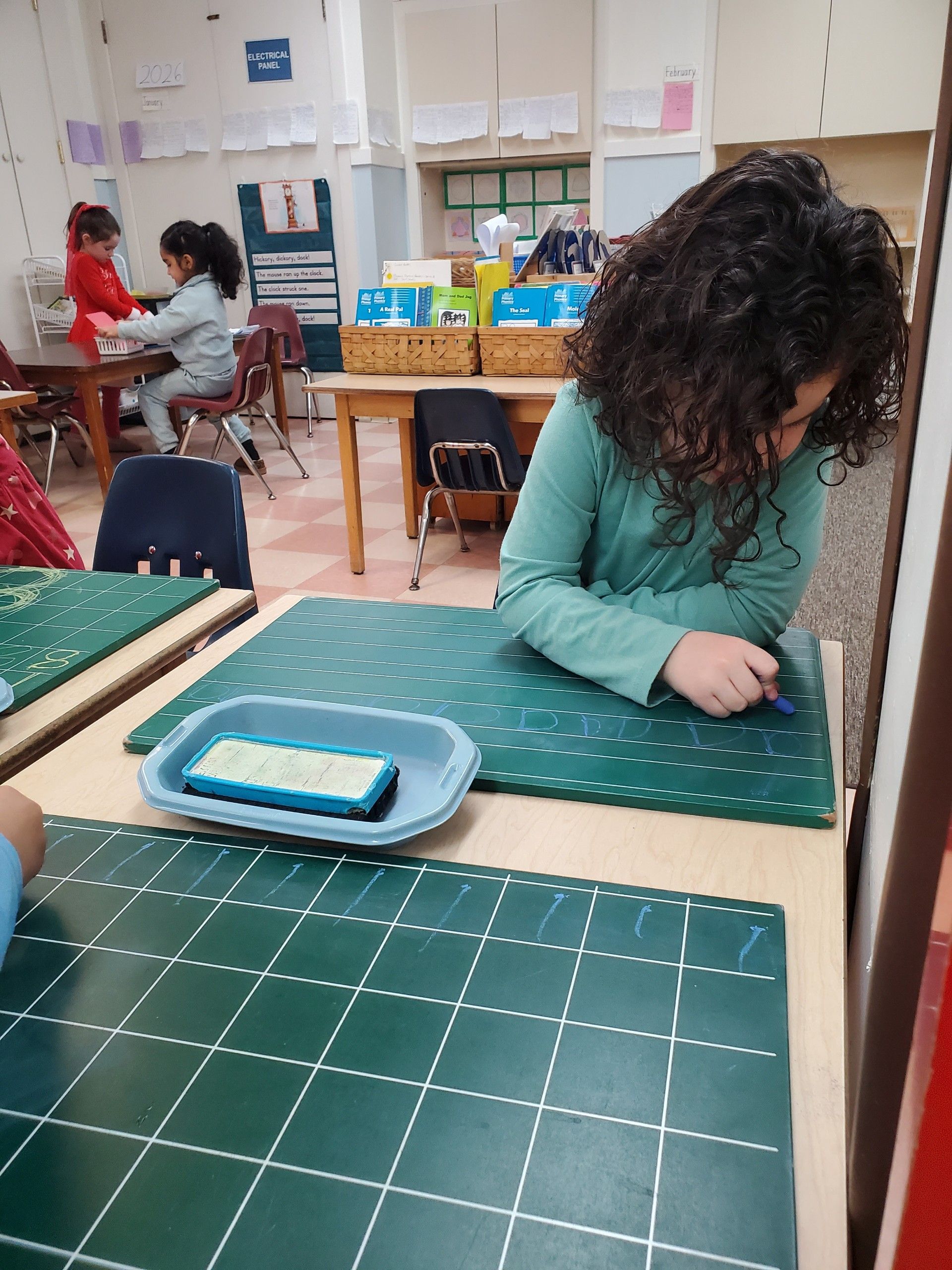 A student works on a green grid mat at a desk in a classroom, with another student visible in the background.