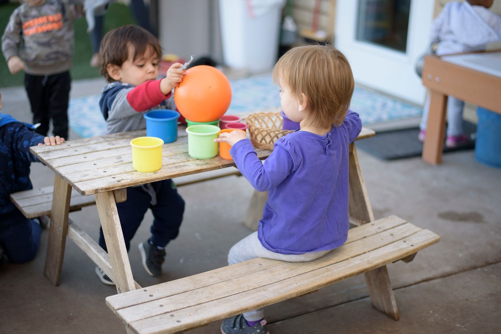Group of children smiling, looking over a white wooden fence outdoors on a sunny day.