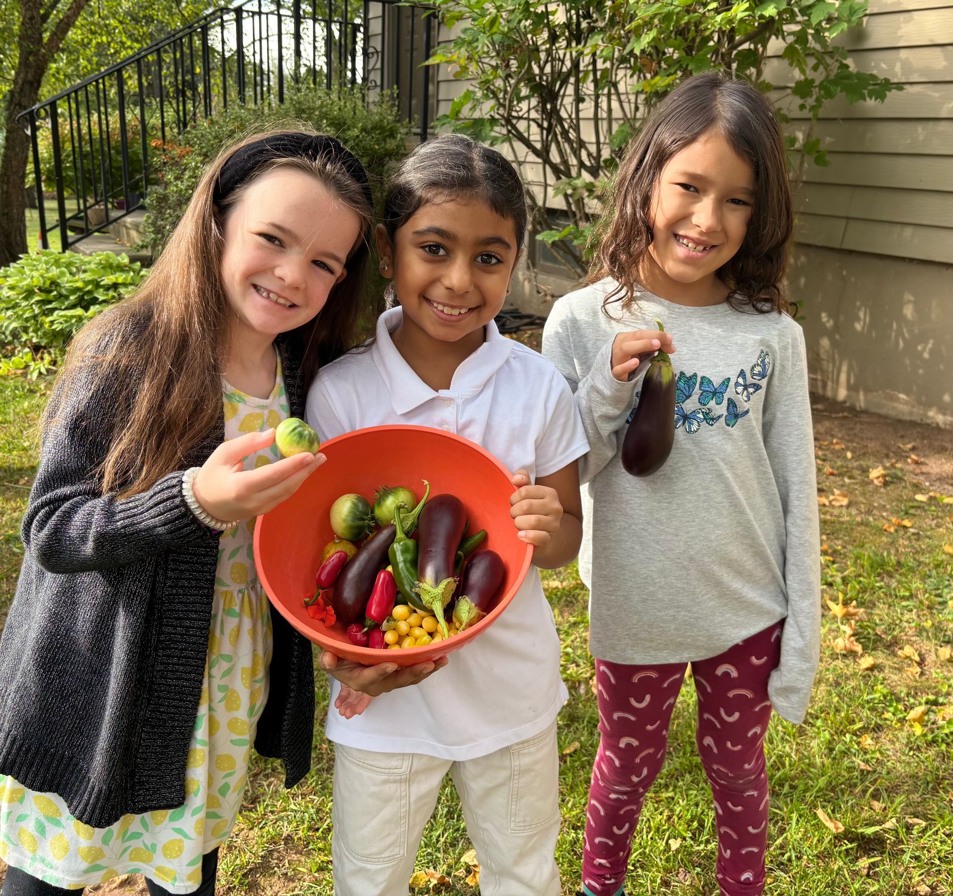 Three children stand outdoors holding a bright orange bowl filled with freshly harvested vegetables.
