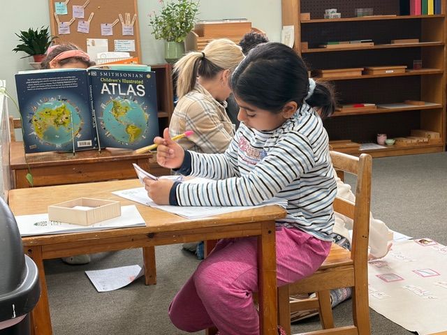 Child writing at a classroom desk beside a bookshelf and atlas display