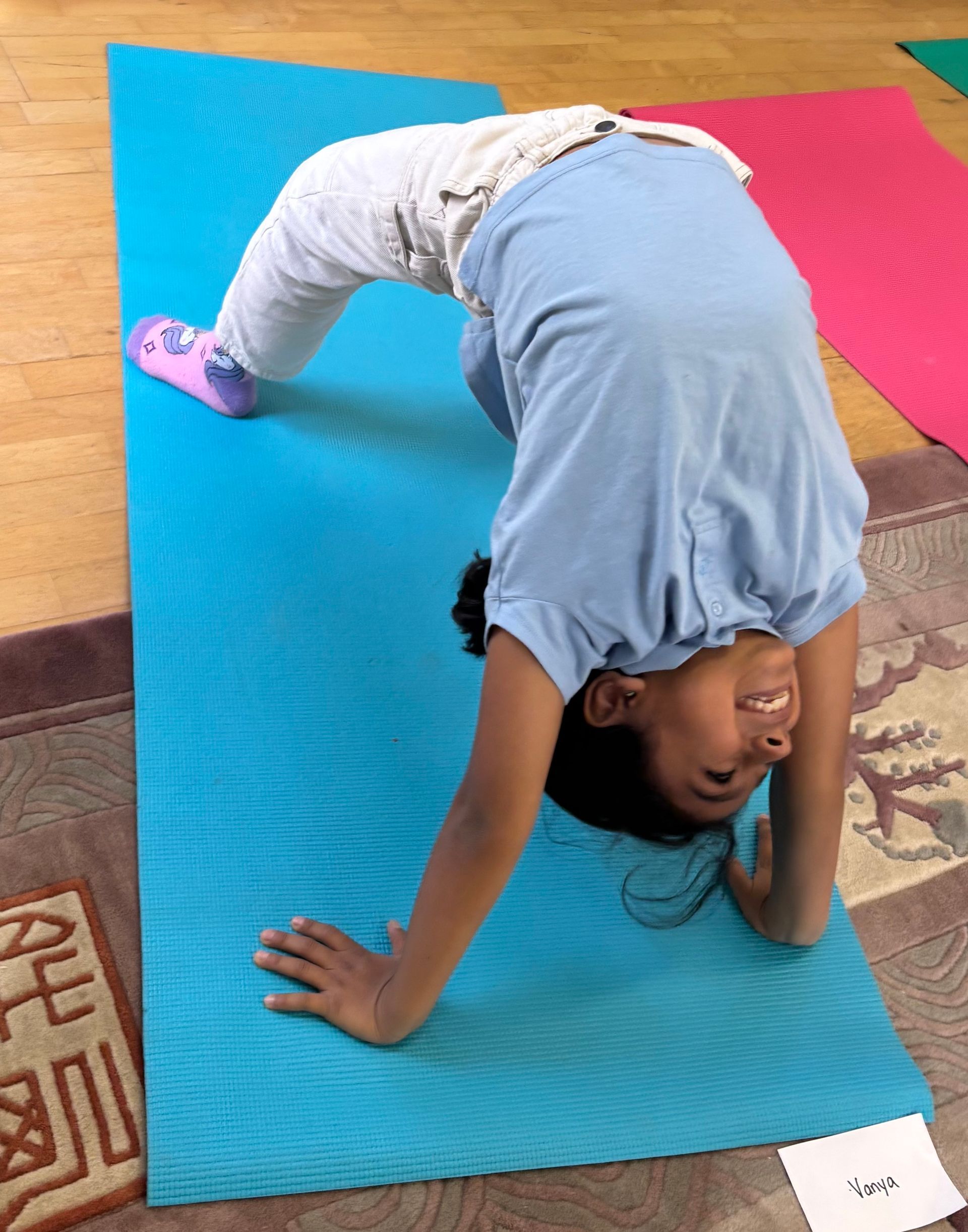 Child doing a backbend yoga pose on a blue mat beside a pink mat indoors