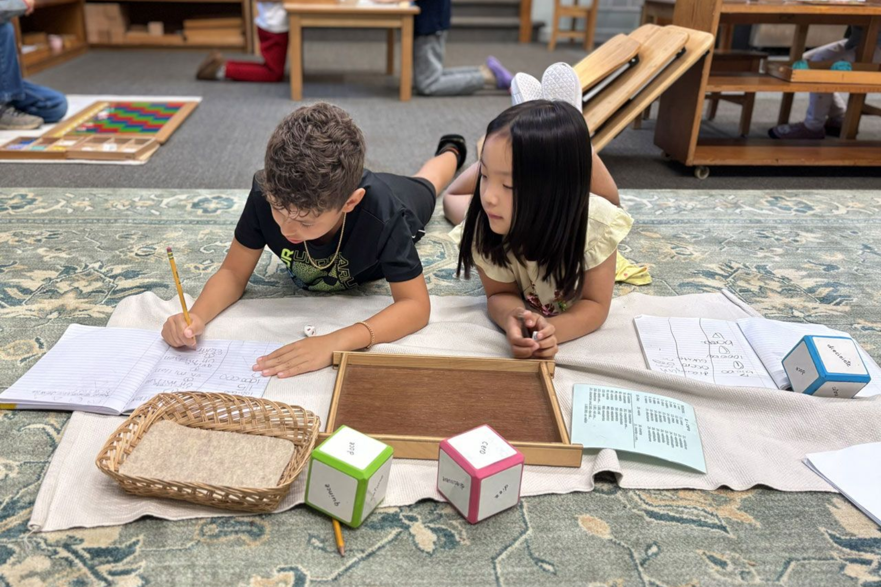 Three Montessori students are sitting on a wooden dock near a lake.