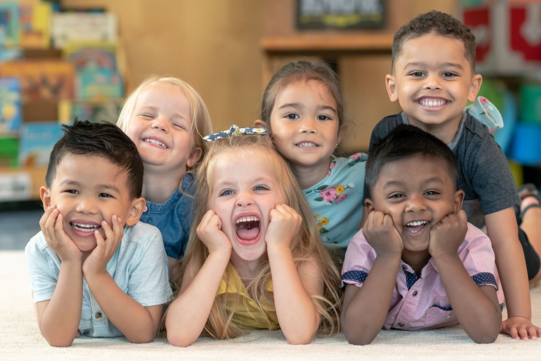 A diverse group of six smiling and laughing toddlers lying together on a classroom rug.