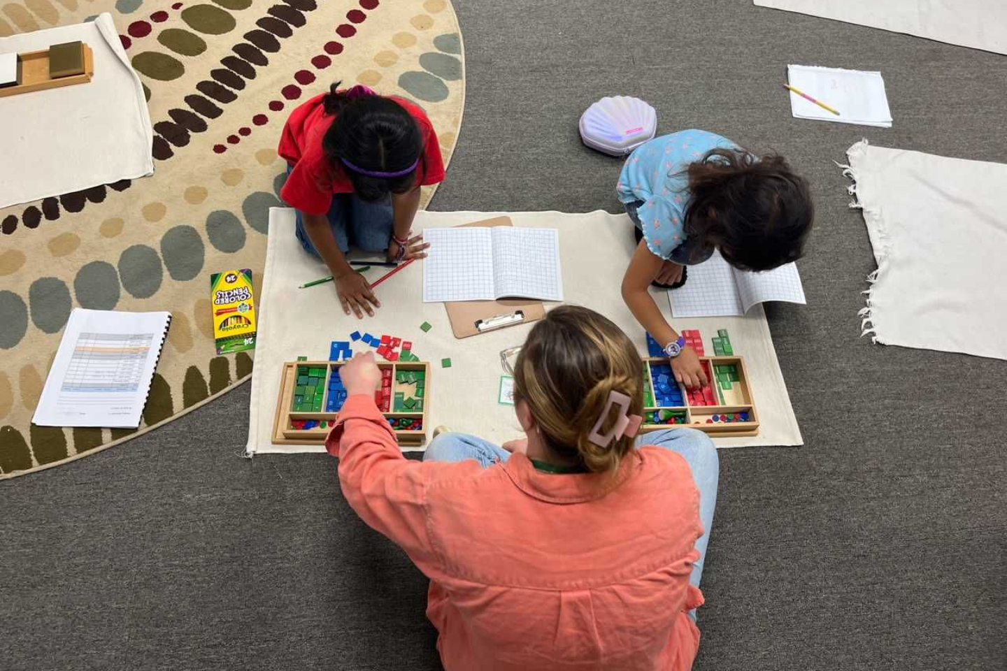 Elementary students using Montessori math materials and the Stamp Game for hands-on learning with their teacher in a classroom setting