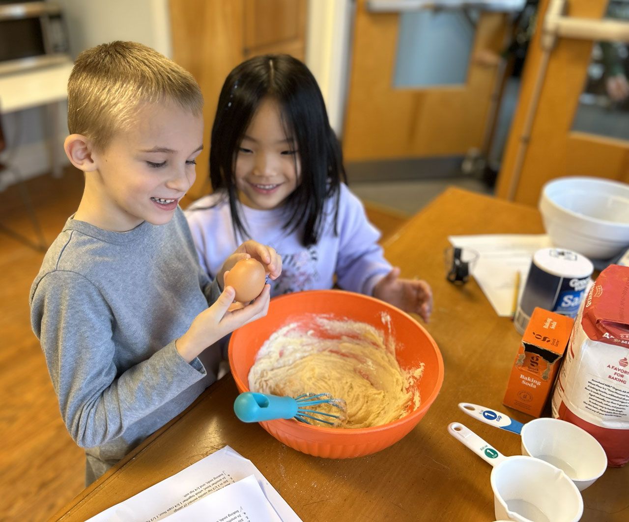 Two Montessori elementary students working together in a kitchen to crack an egg into a bowl of batter for a baking project