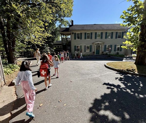 A line of Montessori elementary students walking across a sunny paved driveway toward a gray two-story school building.