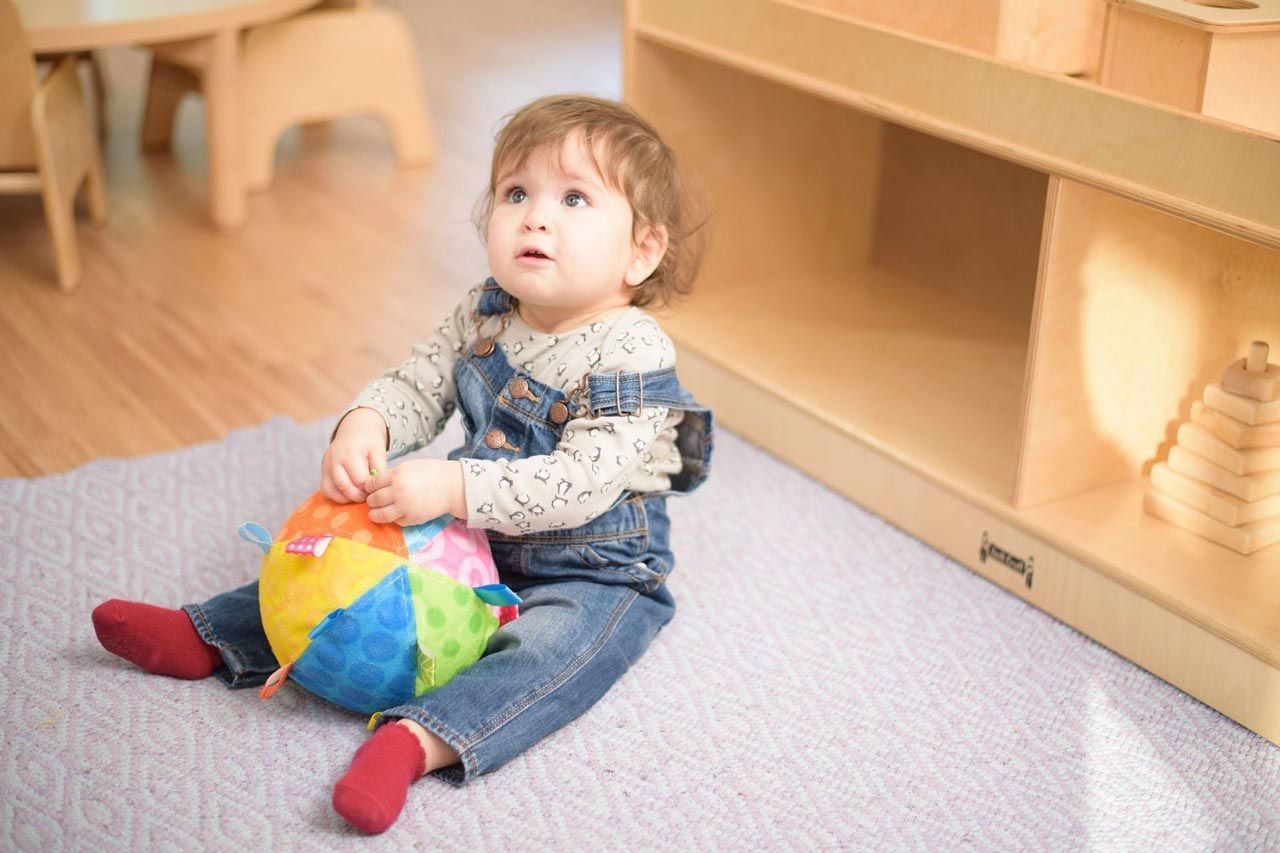 A Montessori Infant is sitting on the floor playing with a ball.