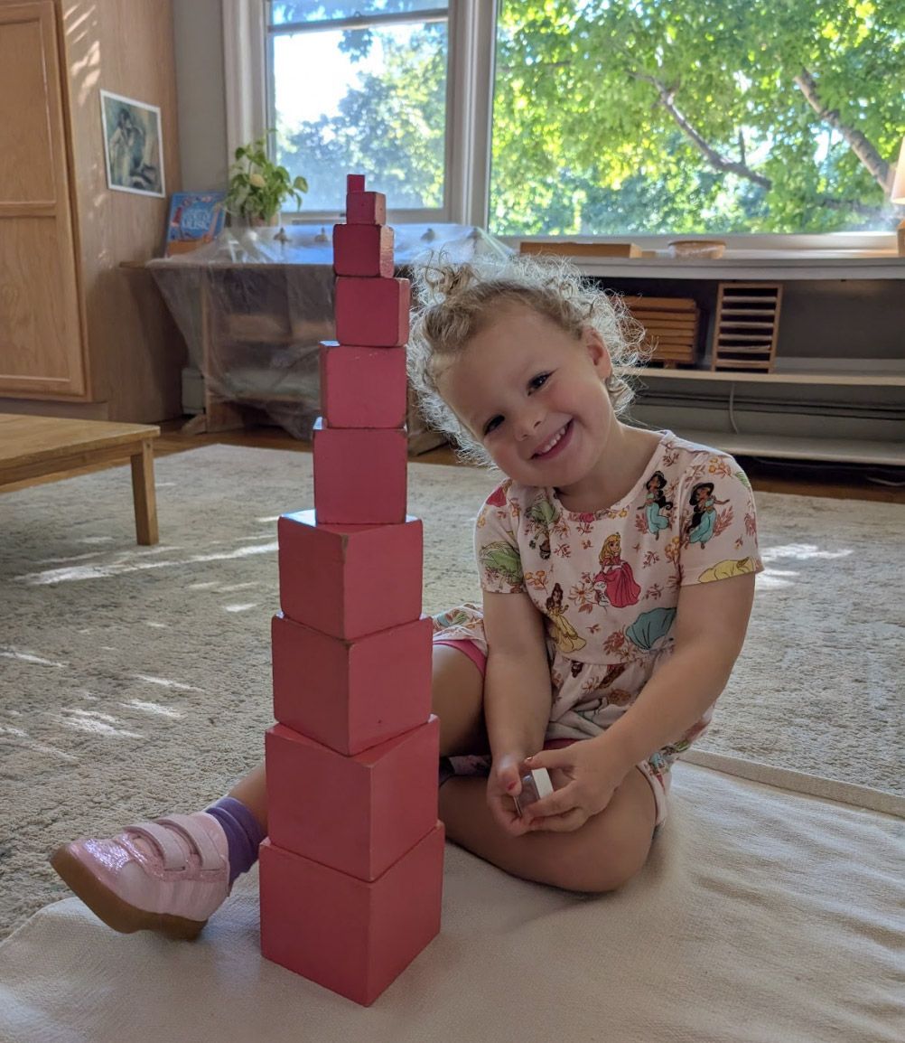A smiling Montessori primary student sitting on a rug next to a fully completed Pink Tower sensorial material in a sunlit classroom.