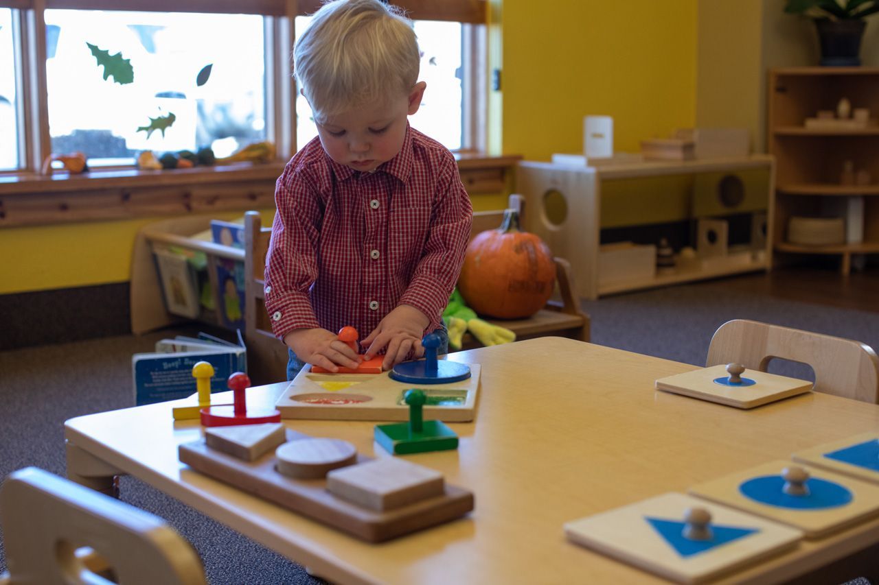 A Montessori toddler standing at a wooden table and focusing on a geometric shape puzzle with large knobs.