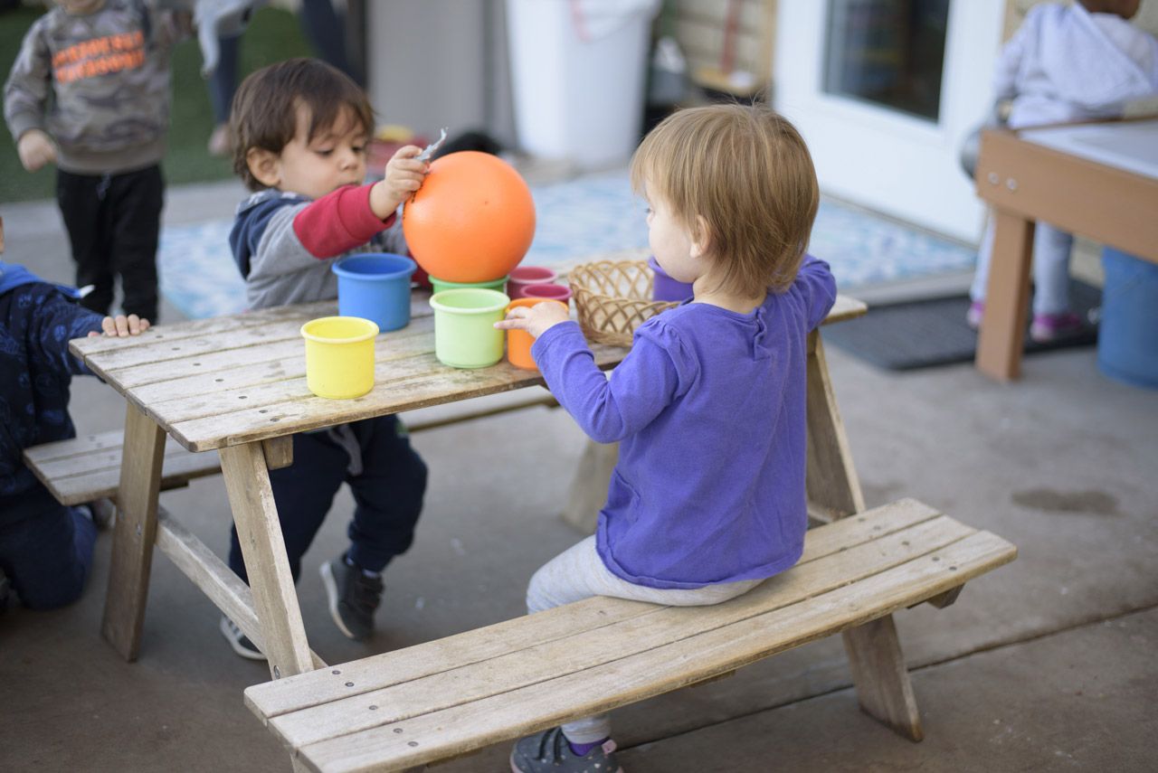 Two Montessori toddlers working together at an outdoor picnic table with colorful stacking cups and an orange ball