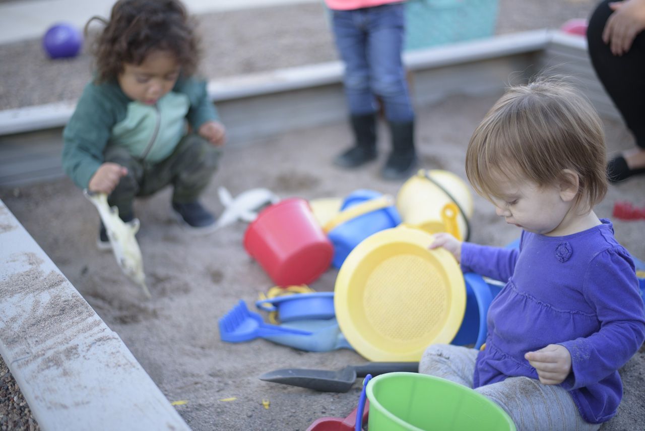 Two Montessori toddlers sitting in an outdoor sandbox playing with colorful buckets, shovels, and sifters.