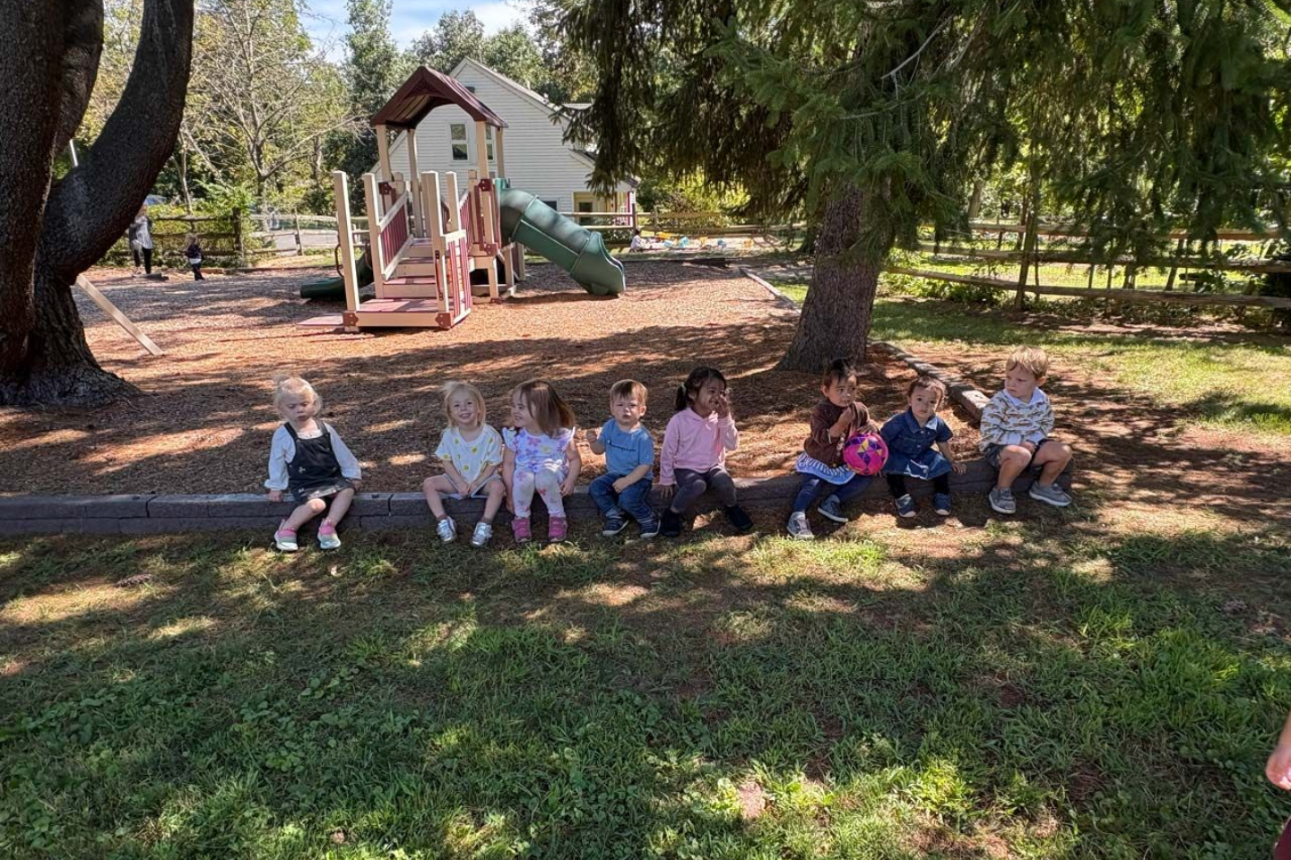Group of Montessori toddlers sitting together in the shade on the playground border