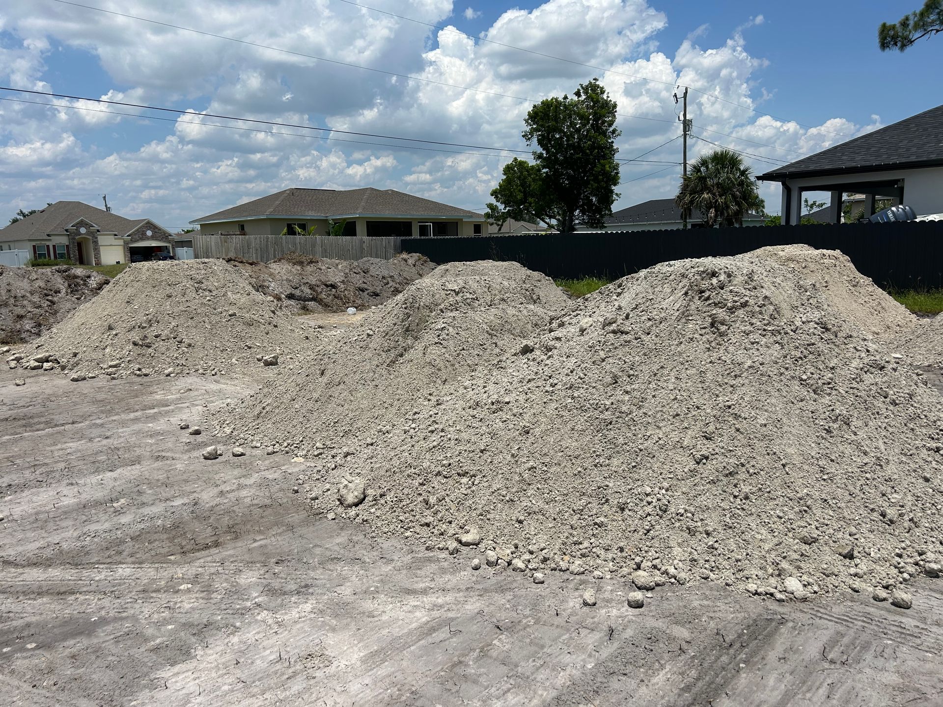 Piles of light-colored gravel on a dusty surface, in front of houses under a cloudy sky.