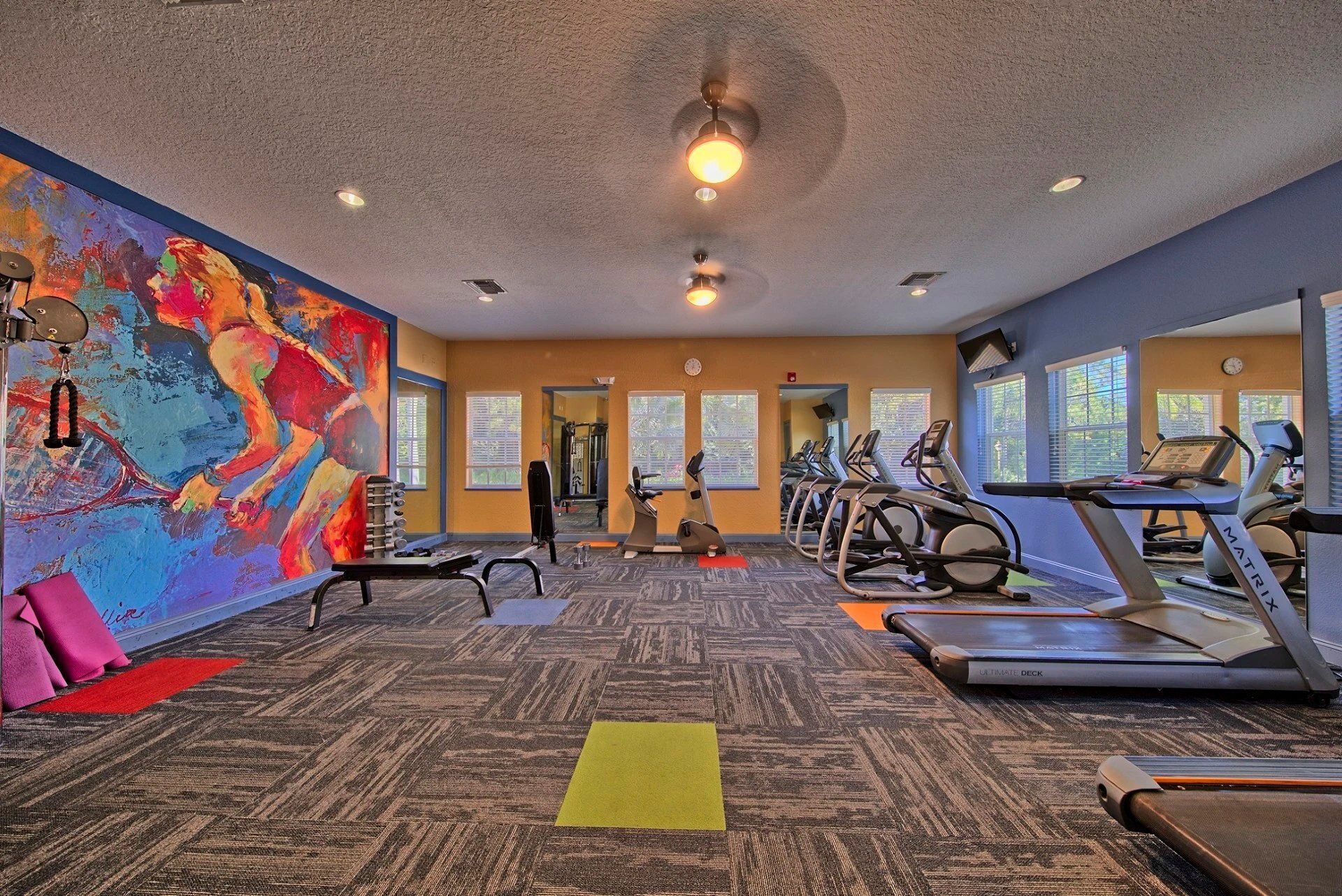 A spacious gym featuring colorful artwork, exercise machines, and workout mats in a well-lit environment at Toledo Club Apartment Homes, offers apartments for rent in North Port, FL.