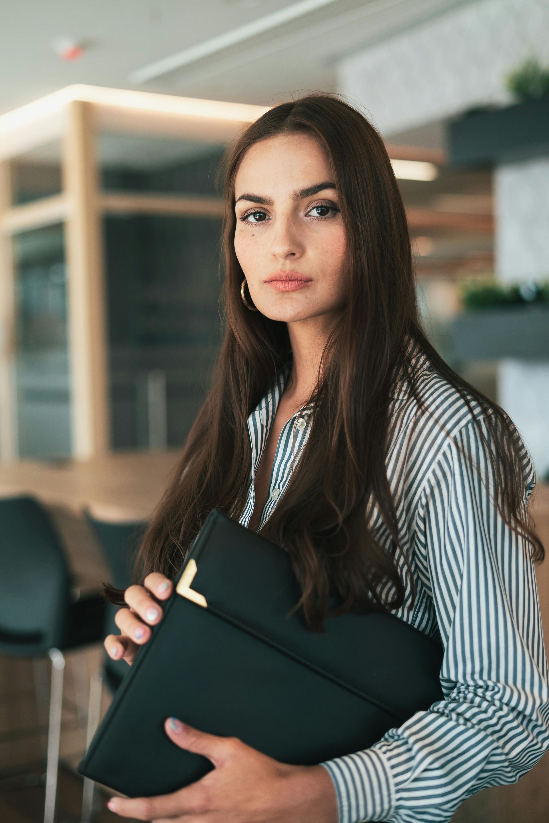 Woman in striped shirt holds a black folder, looking at the camera in an office setting.
