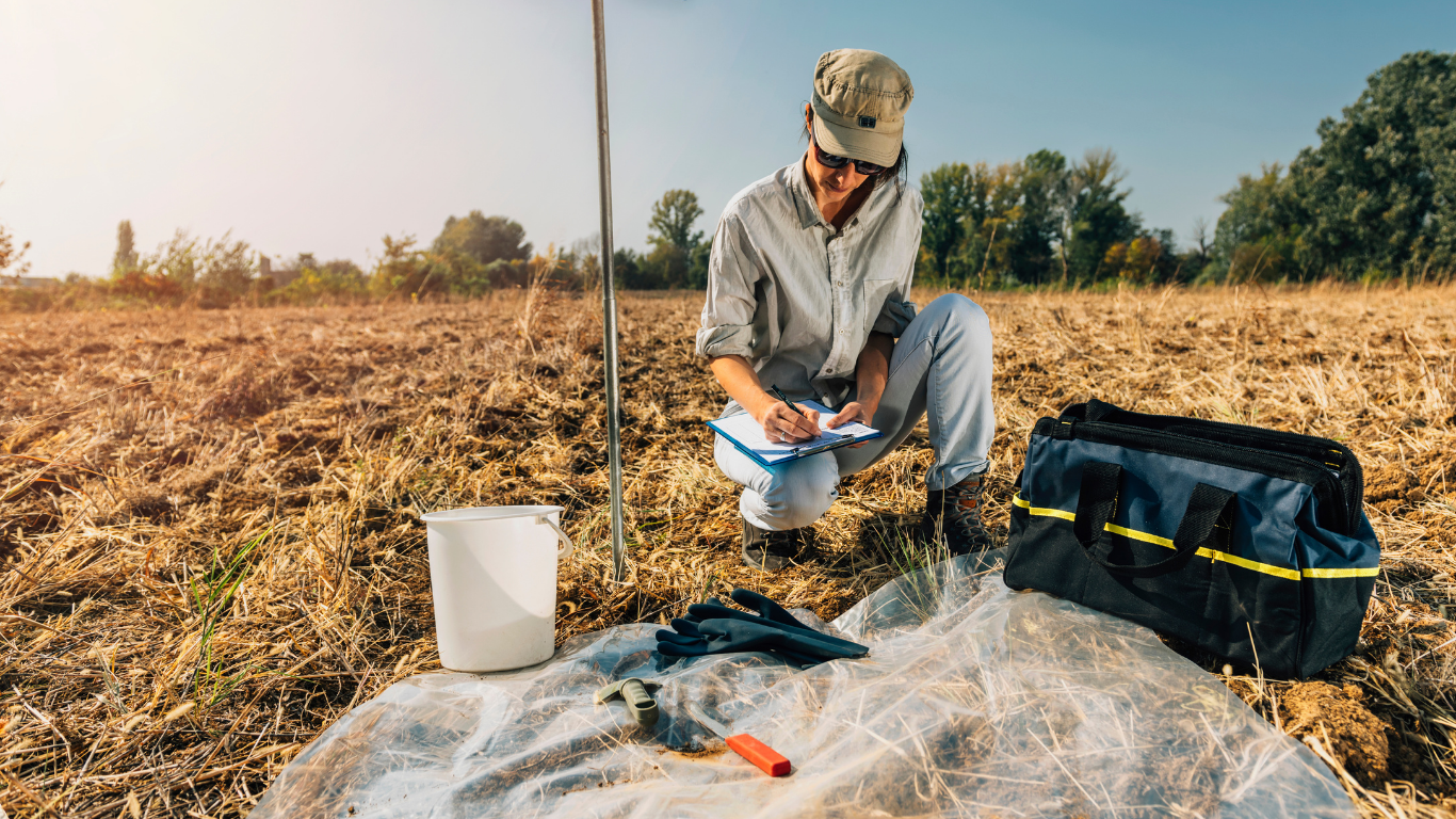 Person taking notes in a field, with soil sampling tools and a bag.