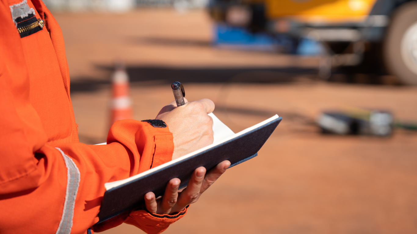 Person in orange workwear writing in a notebook outdoors, blurred background of machinery.
