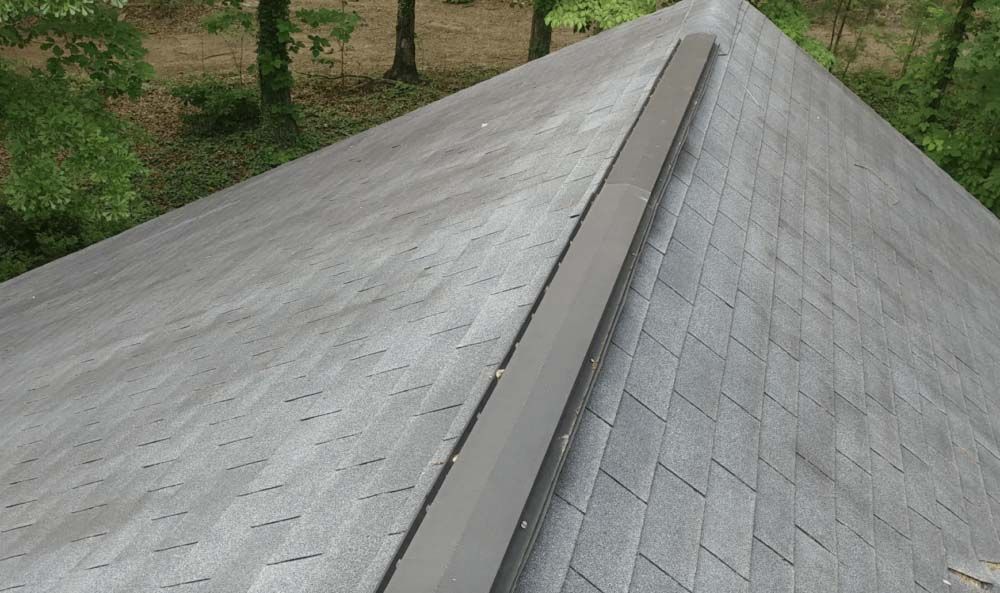 View of a gray, weathered shingled roof with a dark ridge cap, surrounded by trees.