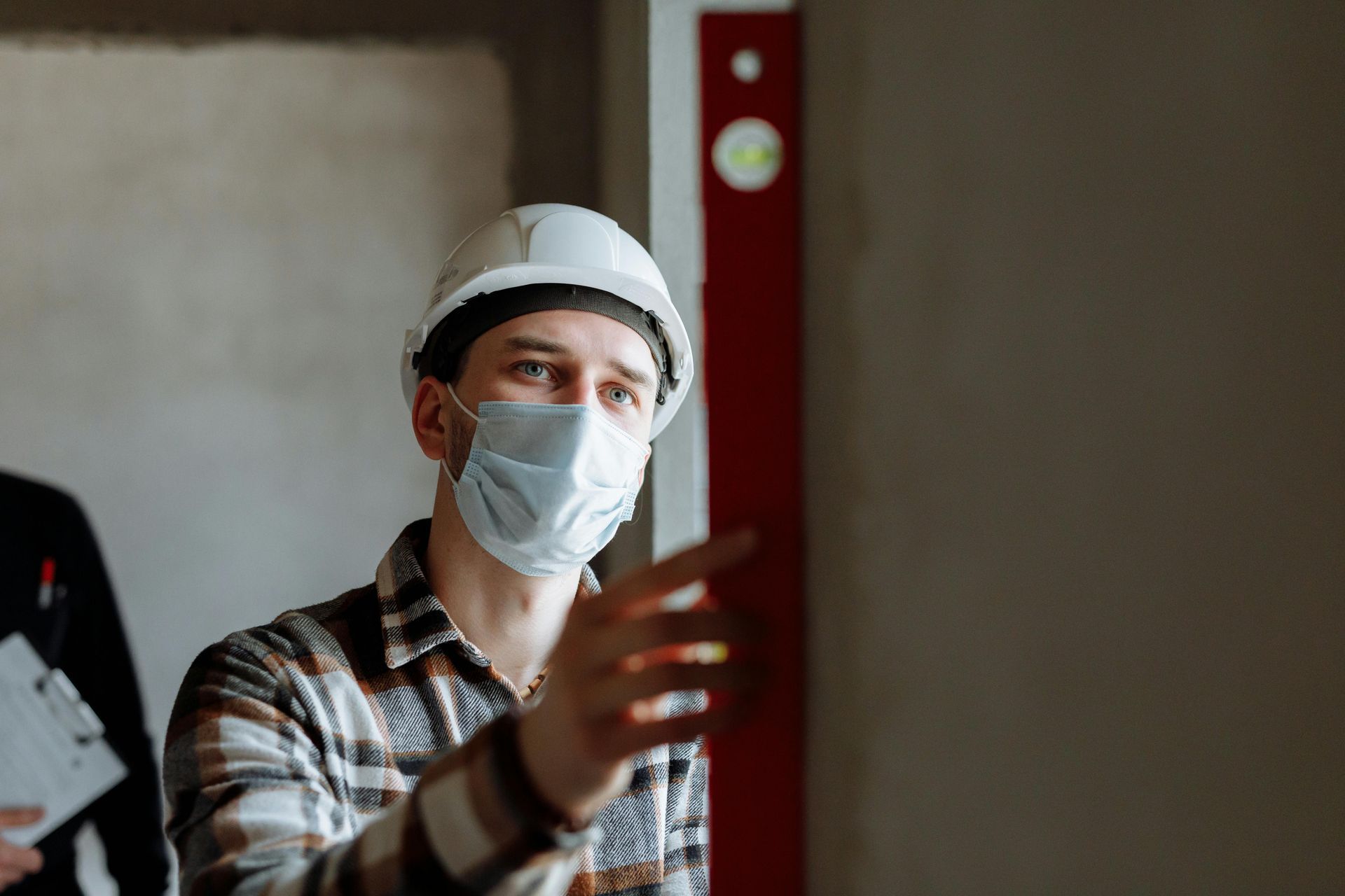 A construction worker in a white hard hat and face mask points toward a wall inside a building under construction.