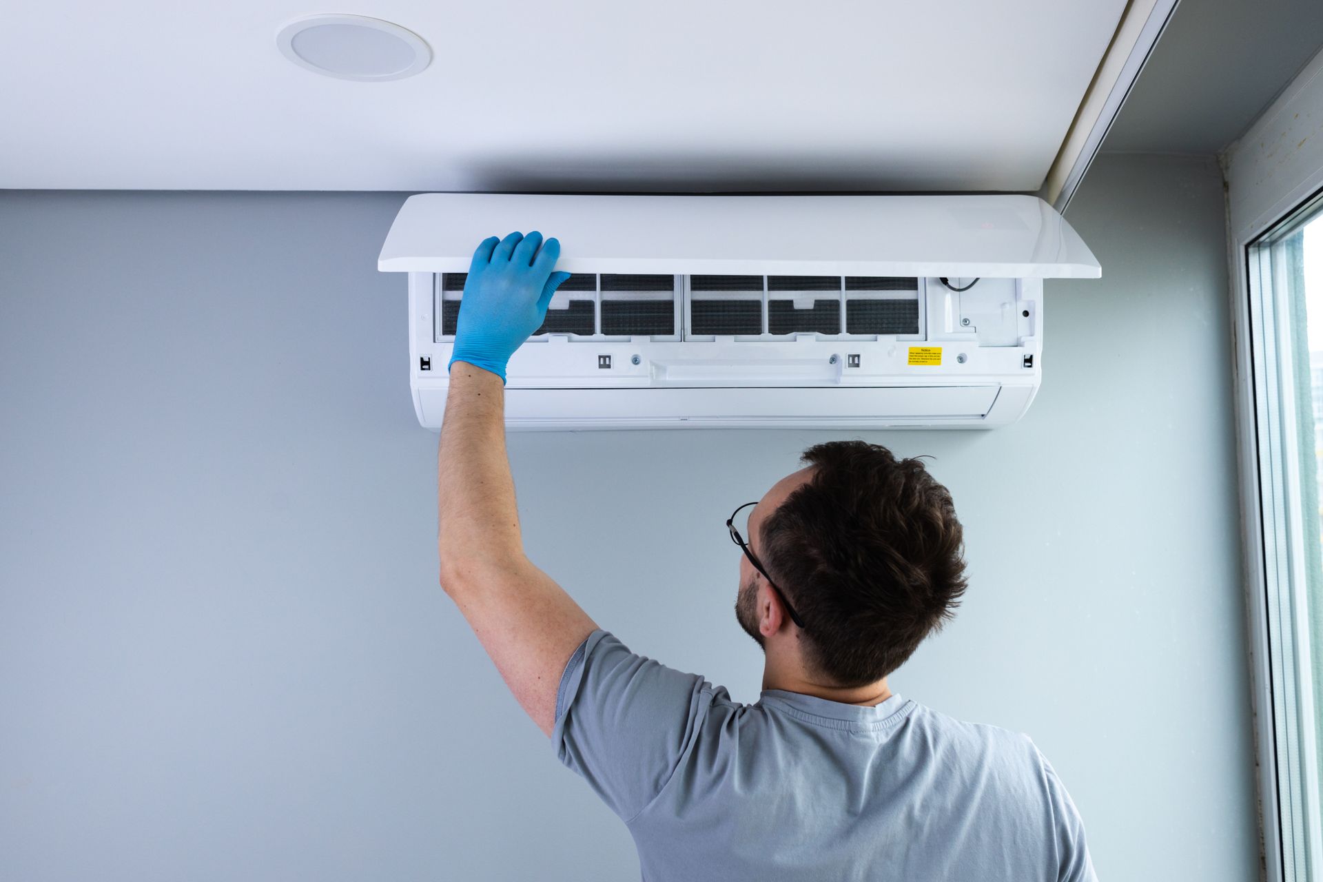 A technician in a gray shirt and blue gloves opens the cover of a wall-mounted air conditioning unit for maintenance.