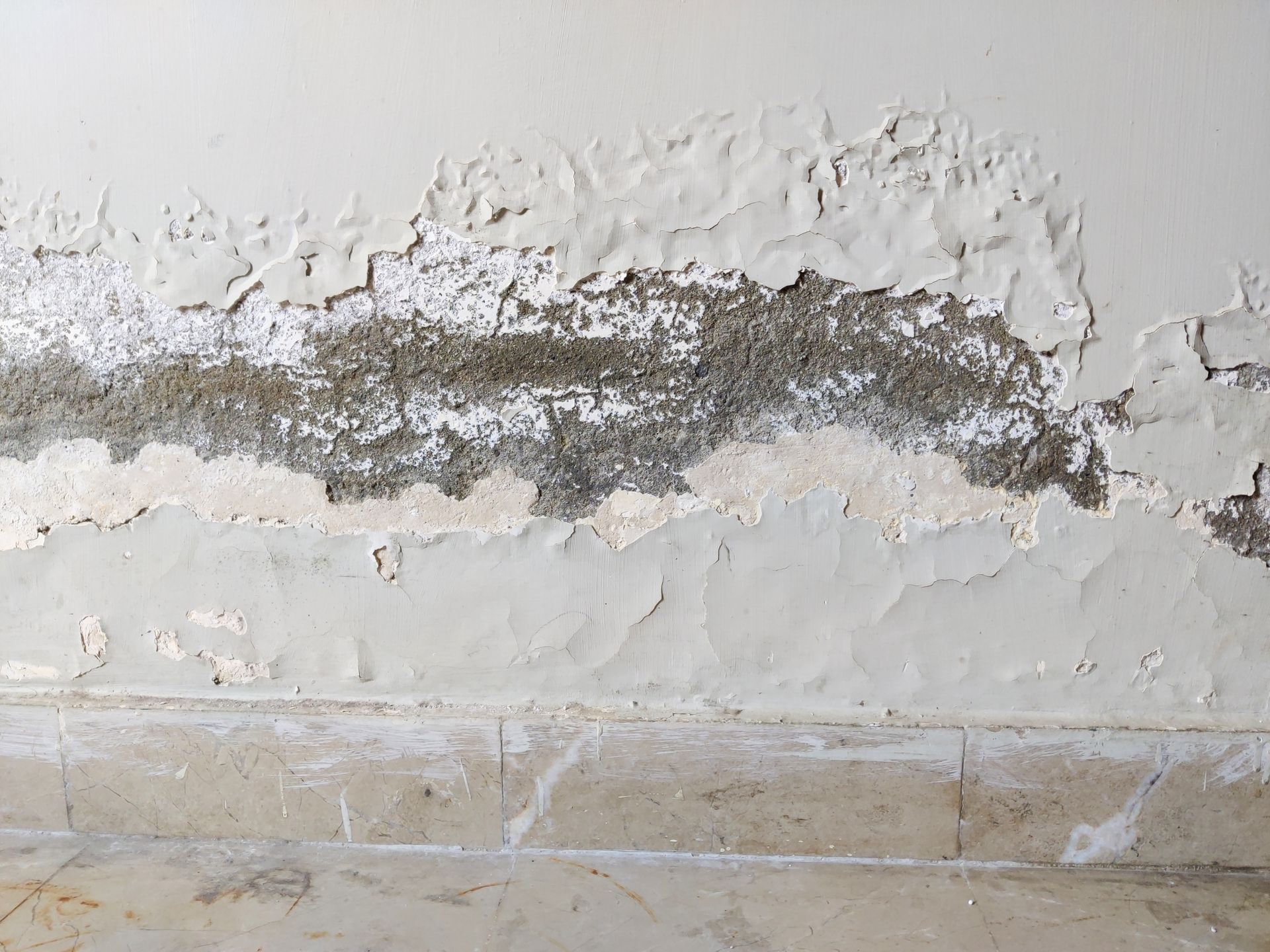 Peeling, damaged white wall paint with visible gray plaster beneath, located above a tiled floor.