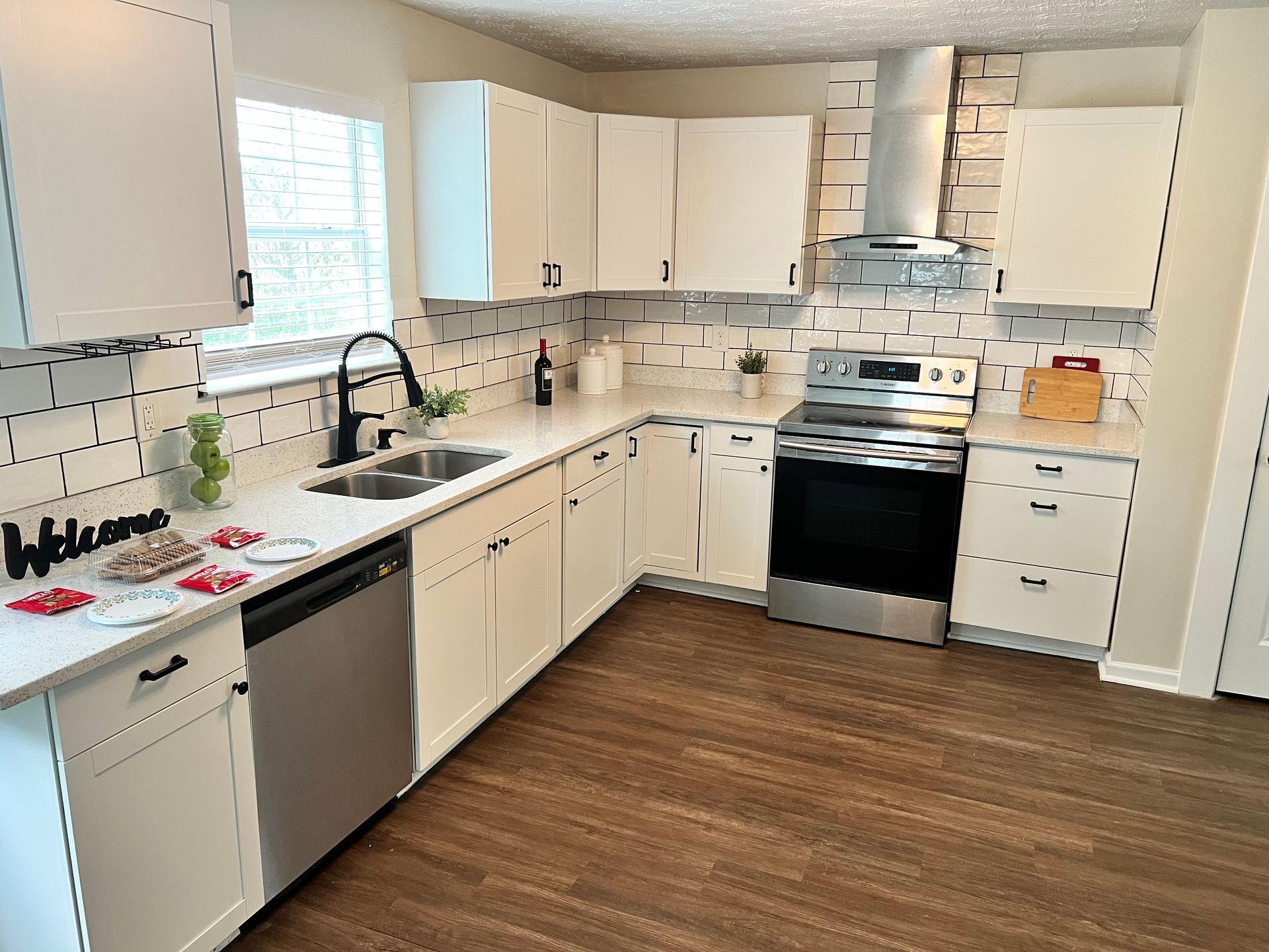Modern white kitchen with stainless steel appliances, dark hardware, and wood-look flooring.