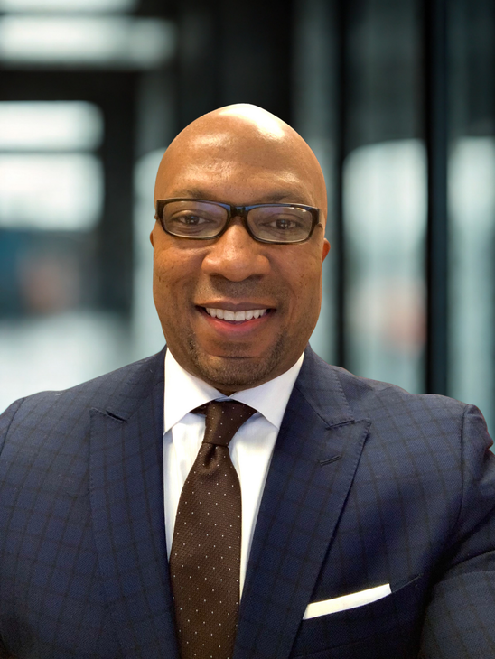 Man in a suit smiles, wearing glasses, against a blurred office background.