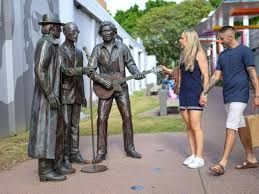 Three bronze statues of musicians beside two people walking on a path outdoors
