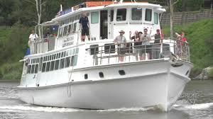 White passenger boat cruising on a river, with people standing on deck and green riverbanks in the background