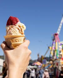 Hand holding a strawberry-topped ice cream cone at a sunny fairground with rides in the background