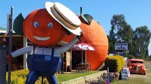 Large roadside orange fruit-shaped statue with a smiling face and hat outside a building