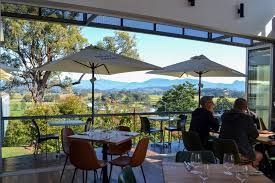 Outdoor cafe terrace with two people seated under umbrellas, overlooking a scenic mountain valley