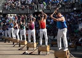 Lumberjacks in a competition, swinging axes to chop logs, outdoors. Crowd in the background.