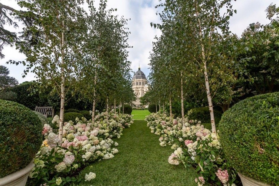 Lined pathway of trees and flowers leading to a large building under a cloudy sky.