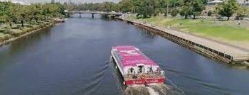 A pink and red boat travels down a wide river flanked by greenery and a long wooden dock, under a bridge.