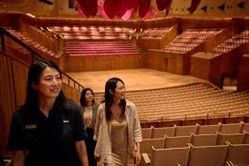 Three women walking in an empty concert hall. Rows of seats, wooden floor, and red lighting.