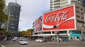 Coca-Cola billboard on a building in a city street, with cars and pedestrians.