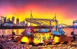 Large outdoor stage with performers lit up by colorful lights at sunset. City skyline in the background.