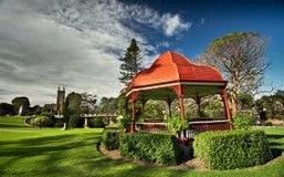 Park scene with red-roofed gazebo, green lawn, trees, and cloudy blue sky.