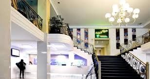 Elegant lobby with grand staircase, chandelier, and a person near a reception desk.