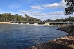 A dam with water flowing over it, trees in the background, blue sky, and a river.