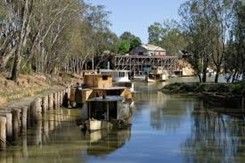 Boats on a river with a wooden structure and buildings in the background. Calm water, sunny day.
