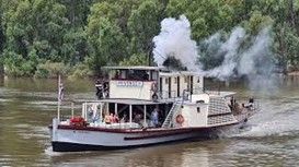 Steam-powered riverboat, white with a black hull, emitting smoke. Passengers onboard, trees in the background.