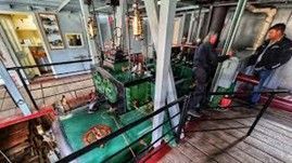 Two men examine a green steam engine in a boat's engine room.