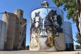 Grain silo with a mural of two horses pulling a wagon; blue sky, sunny day.