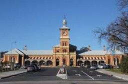 Railway station building with clock tower and arched facade. Cars parked on the road in front.
