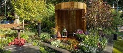 A bamboo-constructed gazebo in a garden. A birdbath sits nearby. Green and red plants surround.